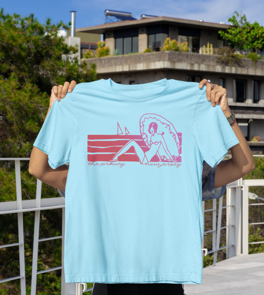 Person holding a light blue t-shirt with a graphic design of a victorian era woman on the beach in a bikini 